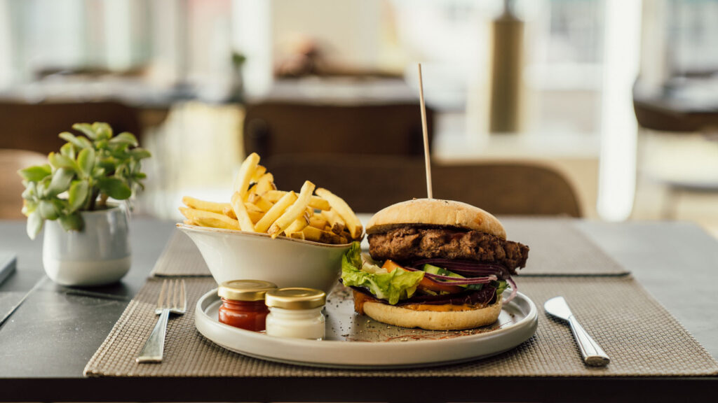 Darstellung eines Resturanttisches mit einem Teller auf dem ein Burger und dazugehörigen Pommes angerichtet sind.